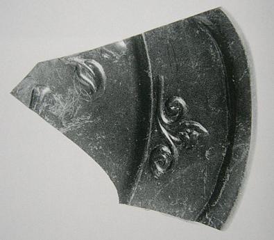 唐草状の浮き彫りが施された黒色の陶器片が欠けた縁と摩耗した表面を示しながら平面上に置かれて質感が際立つ写真