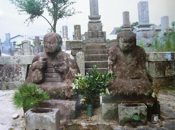 墓地の中央に並ぶ風化した二体の石像が苔むした質感を見せつつ供えられた松や葉の緑に囲まれ静かに佇む写真