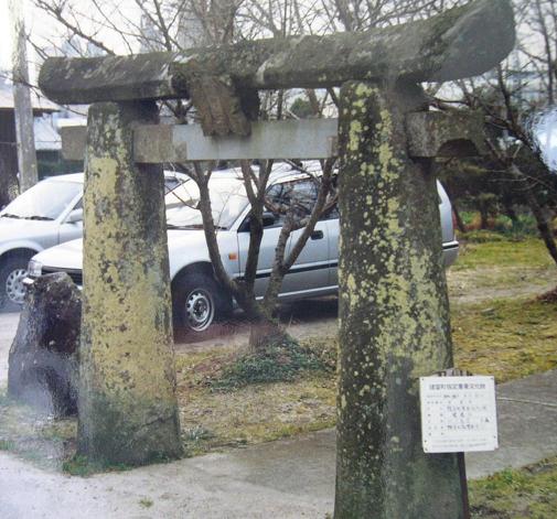 苔むした古い石造鳥居が立ち、周囲の木立や駐車された車と説明板が背景に重なって静かな境内入口の佇まいを示す写真