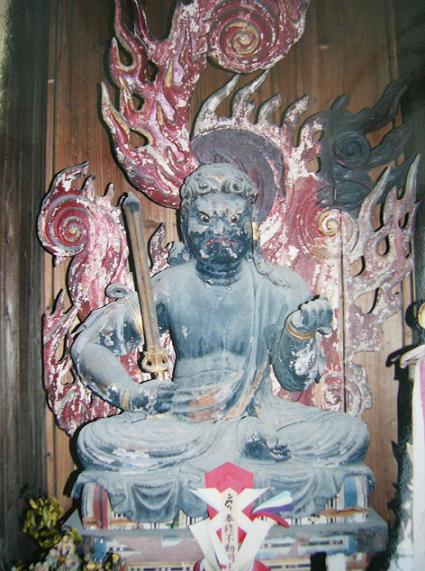 背後に赤い火焔光背が大きく渦巻く古色を帯びた木造の不動明王像が剣と羂索を携え荒々しい表情と重厚な気迫を示して堂内に坐す写真