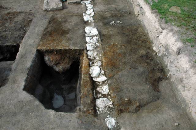 地面が掘り下げられた発掘調査現場において、土層の断面に白い土嚢が縦一列に並んでおり、その横に製鉄の際に発生する鉄滓やレンガなどを捨てた土坑を捉えた写真