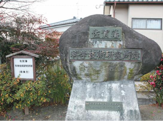 コンクリートの台座に大きな自然石が据えられ、石の表面には「佐賀藩」「科学技術研究所跡」と刻まれたプレートが取り付けられており、左隣には案内板が立つ、史跡の記念碑を正面から捉えた写真