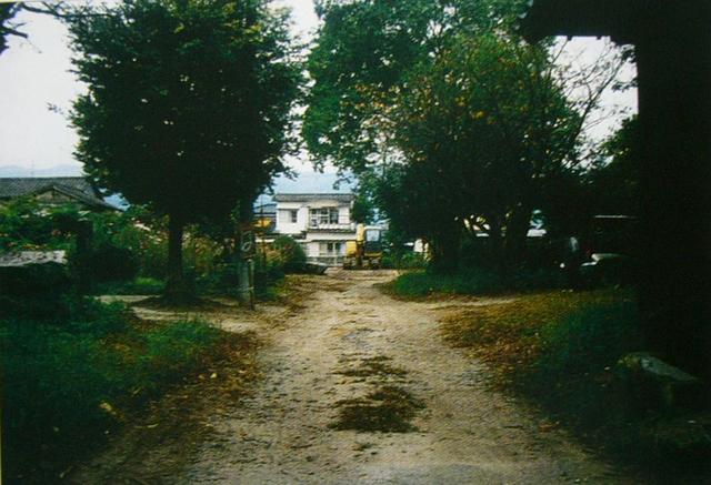 大きな木々に囲まれた未舗装の土の道が奥へと続き、その突き当たりに白い2階建ての住宅と黄色い建設機械のような車両が見える、木漏れ日が差し込む静かな住宅地の一角にある風景を捉えた、肥前国分寺跡講堂付近の写真
