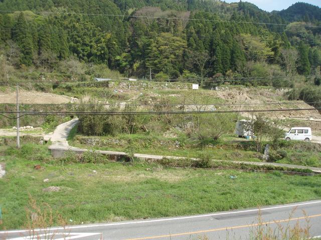 緑豊かな山々を背景に、石垣で区切られた段々畑や空き地が広がるのどかな里山の風景の中を、細いコンクリートの道が蛇行しながら奥へと続き、手前にはアスファルトの道路と白い軽自動車が停車している様子を捉えた、代官所跡の写真