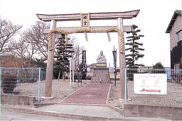 しめ縄が飾られた木造の鳥居の奥に、石造りの台座に鎮座する像へと続く赤い参道があり、周囲をフェンスや整えられた樹木が囲む、燈堂(あかしどう)の写真
