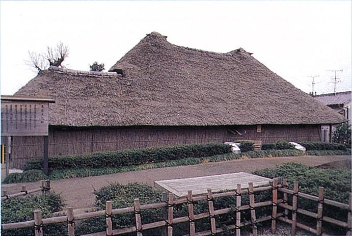竹垣に囲まれた敷地内に建つ、重厚な茅葺き屋根が特徴的な平屋の古民家が、曇り空の下で穏やかな佇まいを見せており、建物の手前には手入れされた低い生垣と未舗装の地面が広がる、歴史を感じさせる山口家住宅の写真