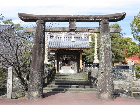 細かな凹凸のある石造りの鳥居が静かに佇み、その奥には瓦屋根の社殿へと続く石段と周囲を囲む豊かな木々が広がっている、石造肥前鳥居を正面から捉えた写真