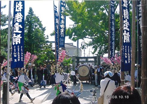 神社で高く掲げられた紺色ののぼり旗が並び、その前で白黒の格子模様の衣装にピンクの花飾りをつけた踊り手が、大きな和太鼓の音に合わせて伝統的な舞を披露している様子の海童神社奉納浮立の写真