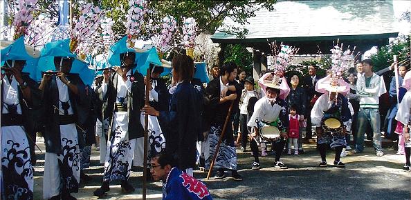 屋外の広場で、波模様が描かれた白い着物や袴をまとい、花飾りがついた大きな笠を被った男性たちが、横笛を吹いたり太鼓を叩いたりしながら舞を披露している松枝神社奉納浮立の写真