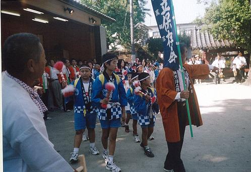 神社の境内で、青いチェック柄の法被を着て赤い飾りのついた棒を持つ子供たちが、のぼり旗を掲げた茶色の法被姿の男性に先導されて列をなし歩いている快万浮立の写真