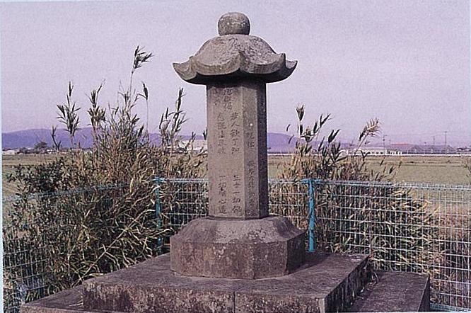 広い田園地帯を背景に文字が刻まれた四角柱の塔身と反り上がる笠を備えた石造塔が風化した質感を見せ静かに佇む写真