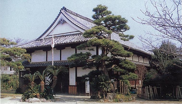 瓦葺きの大きな入母屋屋根を持つ重厚な木造建築の前で、手入れの行き届いた立派な松の木や南国風のソテツが植えられ、白壁と黒い柱をもつ旧佐賀城本丸御座間・堪忍所の外観写真
