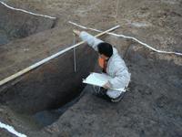 屋外の発掘調査現場において、白い作業服を着た調査員が深く掘削された遺構の縁に屈み、手に持った記録用紙と見比べながら、垂直に立てたスケールを用いて断面の深さや形状を精緻に計測している、遺跡の記録の写真。