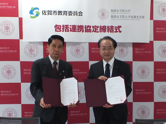 赤と白の市松模様の背景に大学のロゴが並び、中央の横断幕に「包括連携協定締結式」と記された前で、二人の男性が協定書を広げて並んで立っている写真