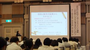 講演会場で多数の参加者がスクリーンに映された「国語と数学の授業をつなぐために」というタイトルのスライドを見ており、壇上には発表者が立っている様子を示す写真