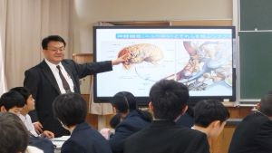 教室内で複数の生徒が机に向かって座り、前方の大型モニターに表示された脳の断面図を指し示す人物が説明している様子を捉えた写真