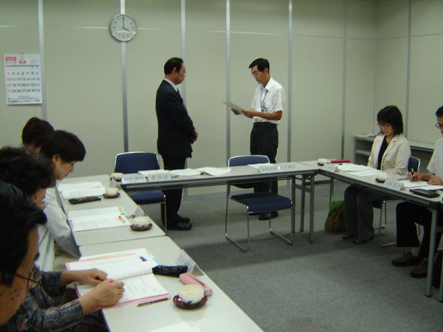 会議室で複数の人々が長机を囲んで書類を広げ、中央では左の男性に向かって右側の男性が文書を読み上げている様子の写真