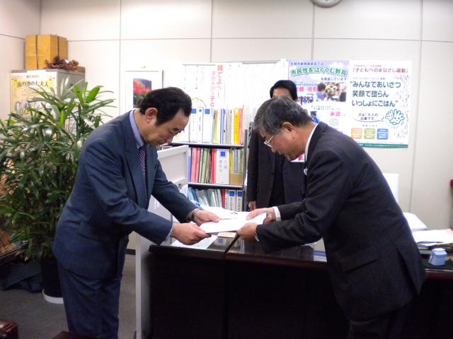オフィス内でスーツ姿の人物が机越しに書類を丁寧に受け渡している写真