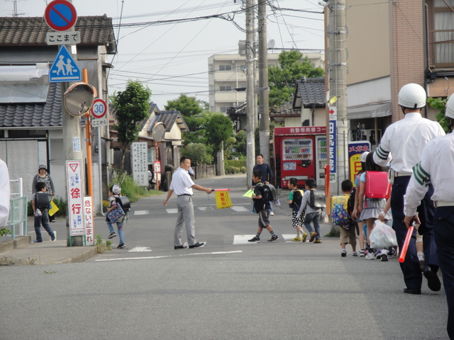 住宅街の横断歩道で、交通誘導員と警察官が立ち、複数の子どもと保護者が道路を横断し、周囲に道路標識と電柱がある通学路安全指導の風景写真