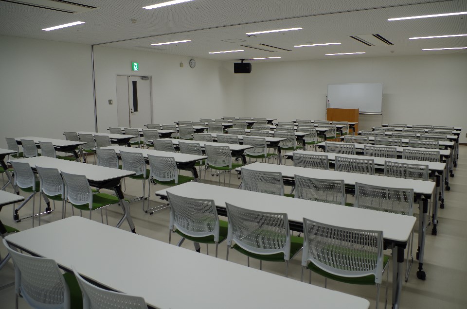 白い壁と天井に囲まれた広い教室で、整然と並ぶ机と椅子が前方の白板と演台へ向かって配置され明るい照明が室内を均一に照らす大会議室の写真