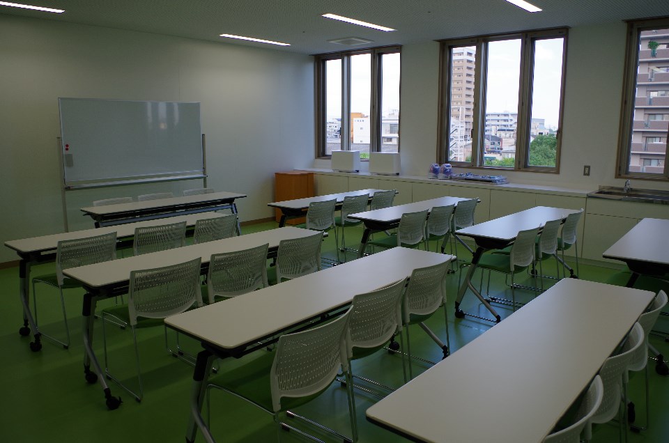 大きな窓から街並みが見える緑色の床の教室で、複数の白い机と椅子が白板と演台へ向け整然と配置された中会議室の写真