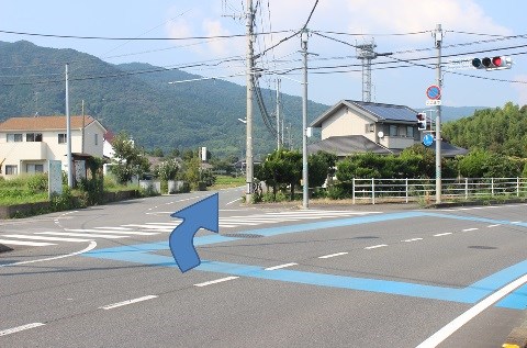 山を背景にした住宅街の交差点で、青色の路面標示がある場所から左前方の道へ進むよう青い曲がり矢印で示した案内写真