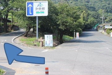 「弘学館 中学校・高等学校」と書かれた看板がある分岐路で、左側の坂道へ進むよう大きな青い矢印で示した案内写真
