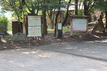 細い木々に囲まれた入口に案内板と地図板が並び背後に木造施設の外観が見える写真
