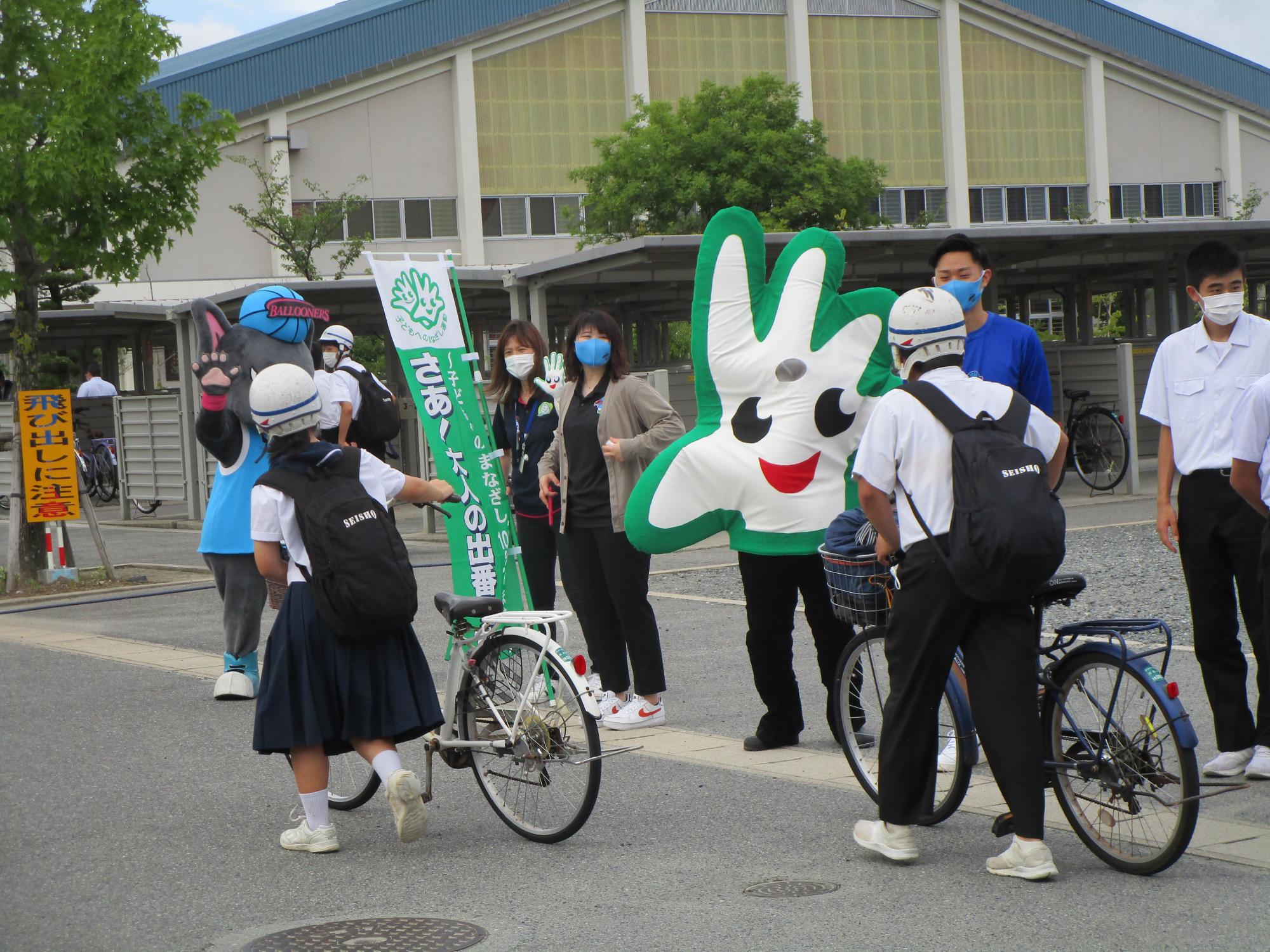 学校の校門前でヘルメットを被った自転車通学の生徒たちが緑のキャラクターやスタッフに迎えられ元気に挨拶を交わしている写真