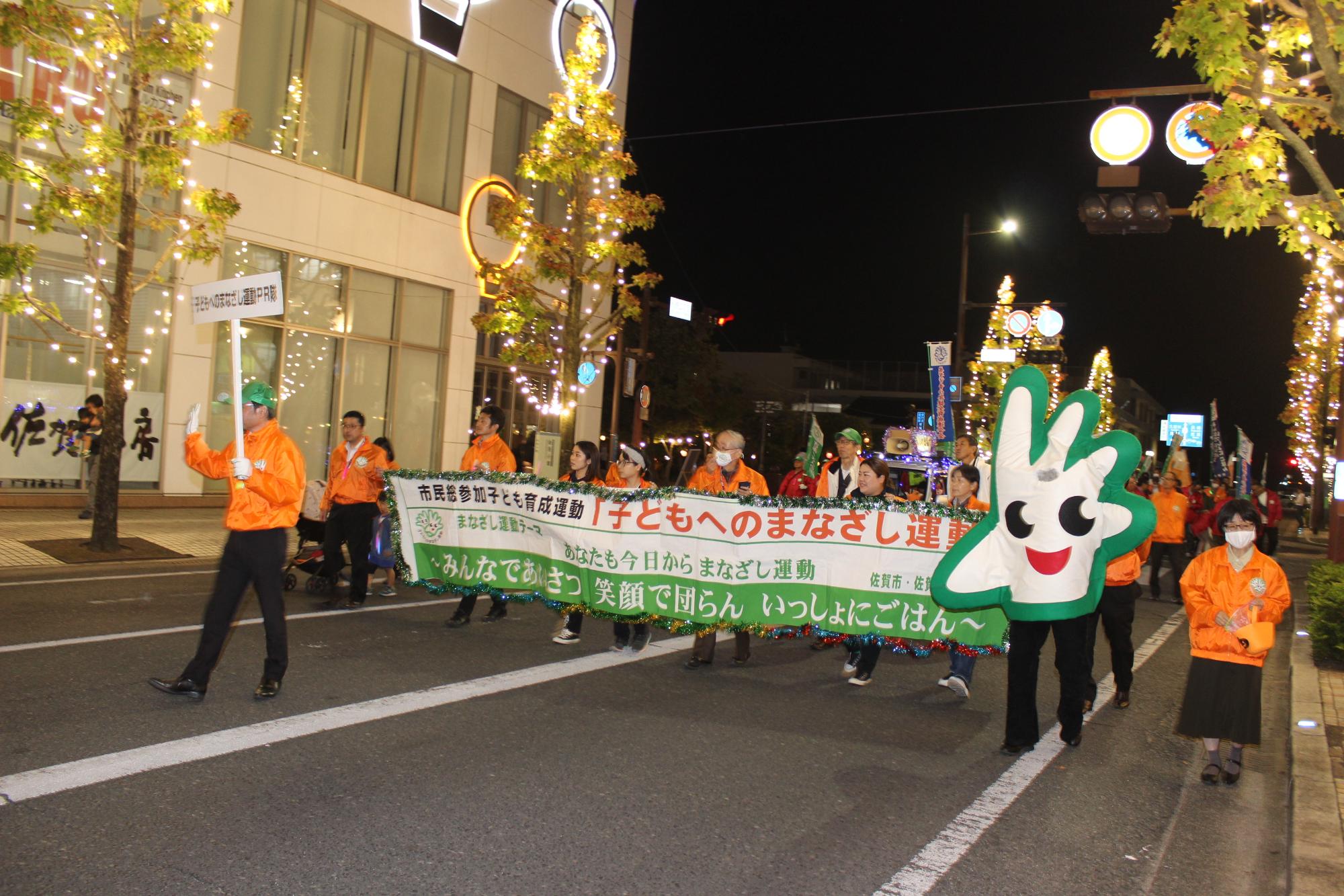 夜の街路で橙色の上着を着た人々と緑のキャラクターが横断幕を掲げながら安全を呼びかけて行進する様子の写真