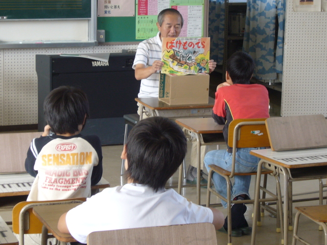 教室で大人が机上の箱を使って紙芝居を掲げ児童たちが前方で静かに聞き入る読み聞かせの様子の写真
