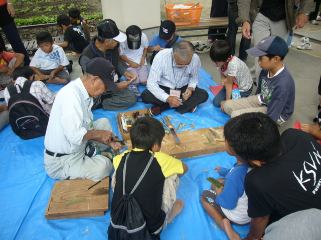 屋外の青いシート上で大人と子どもが囲んで竹トンボ作りの作業に熱心に取り組む賑やかな体験風景の写真