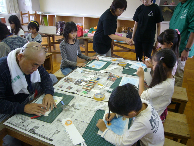 教室内の机を囲んだ子どもと大人が紙や筆や道具を使って熱心にパステル画に取り組む様子の写真
