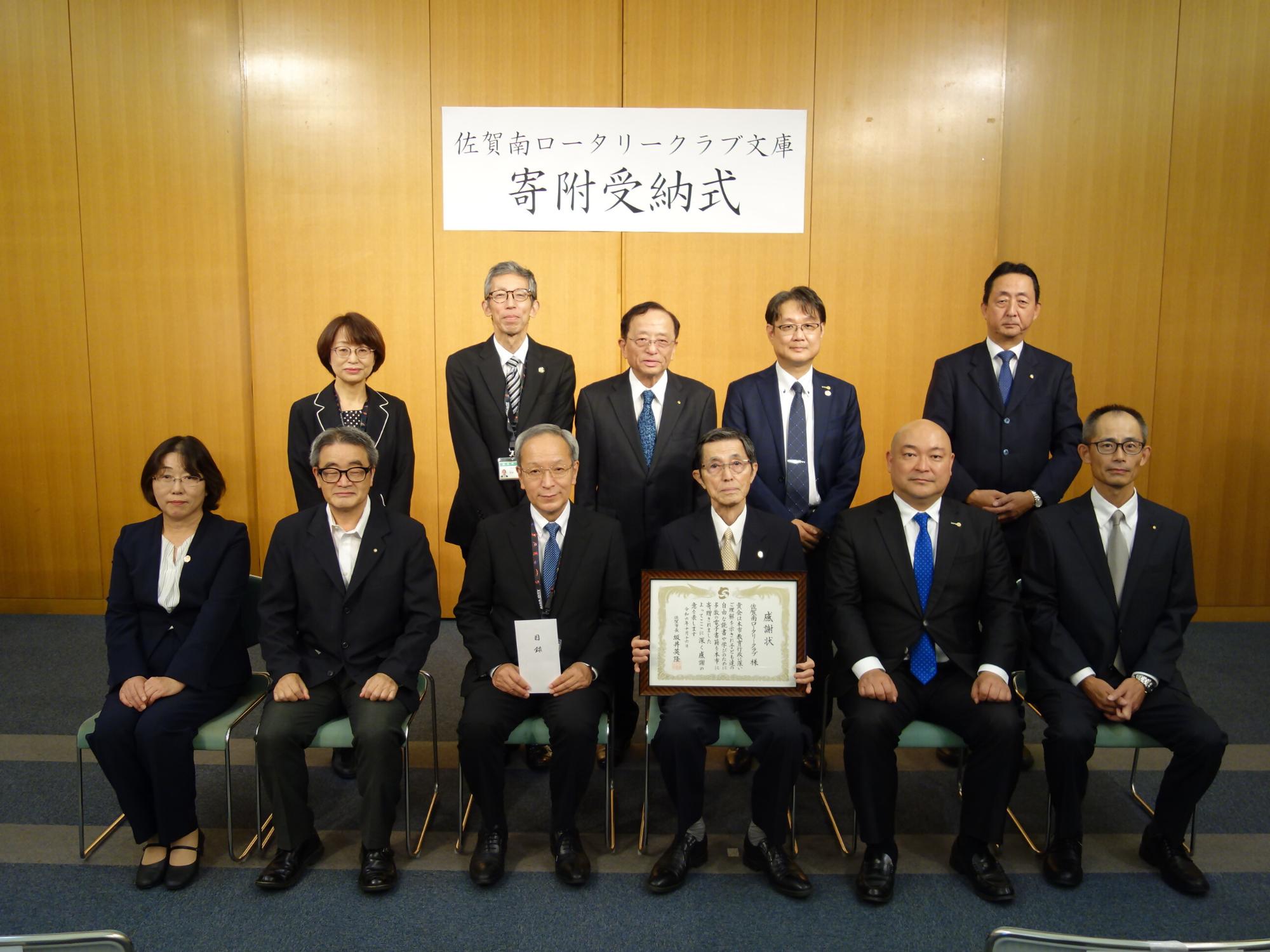 木目調の壁に「佐賀南ロータリークラブ文庫 寄附受納式」の案内板が掲げられ、スーツ姿の関係者達が2列に並び2人が感謝状や目録を持ち記念撮影する写真