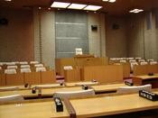 正面奥には、議長席とみられる一段高くなった演壇があり、演壇に向かって、 横長の机と椅子が整然と列をなして並んでいる議場を写した写真