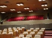 議員席の木製デスクと白い椅子が並び、その奥の一段高い場所に赤い背もたれの傍聴席が設置された演壇から写した写真