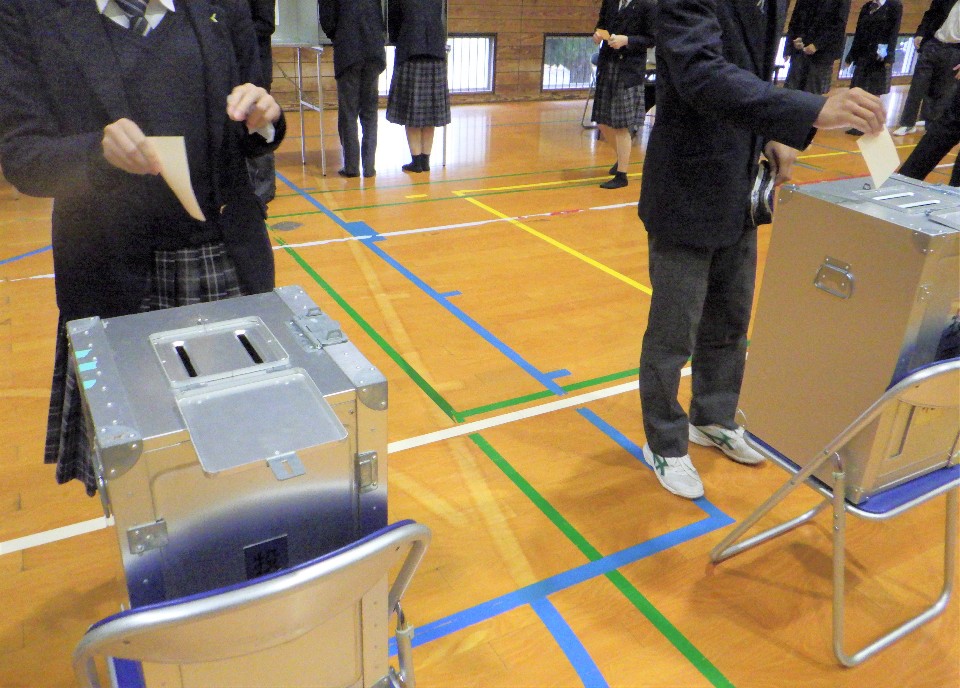 体育館で制服を着た生徒たちが、銀色の投票箱に白い用紙を投函している様子を捉えた写真