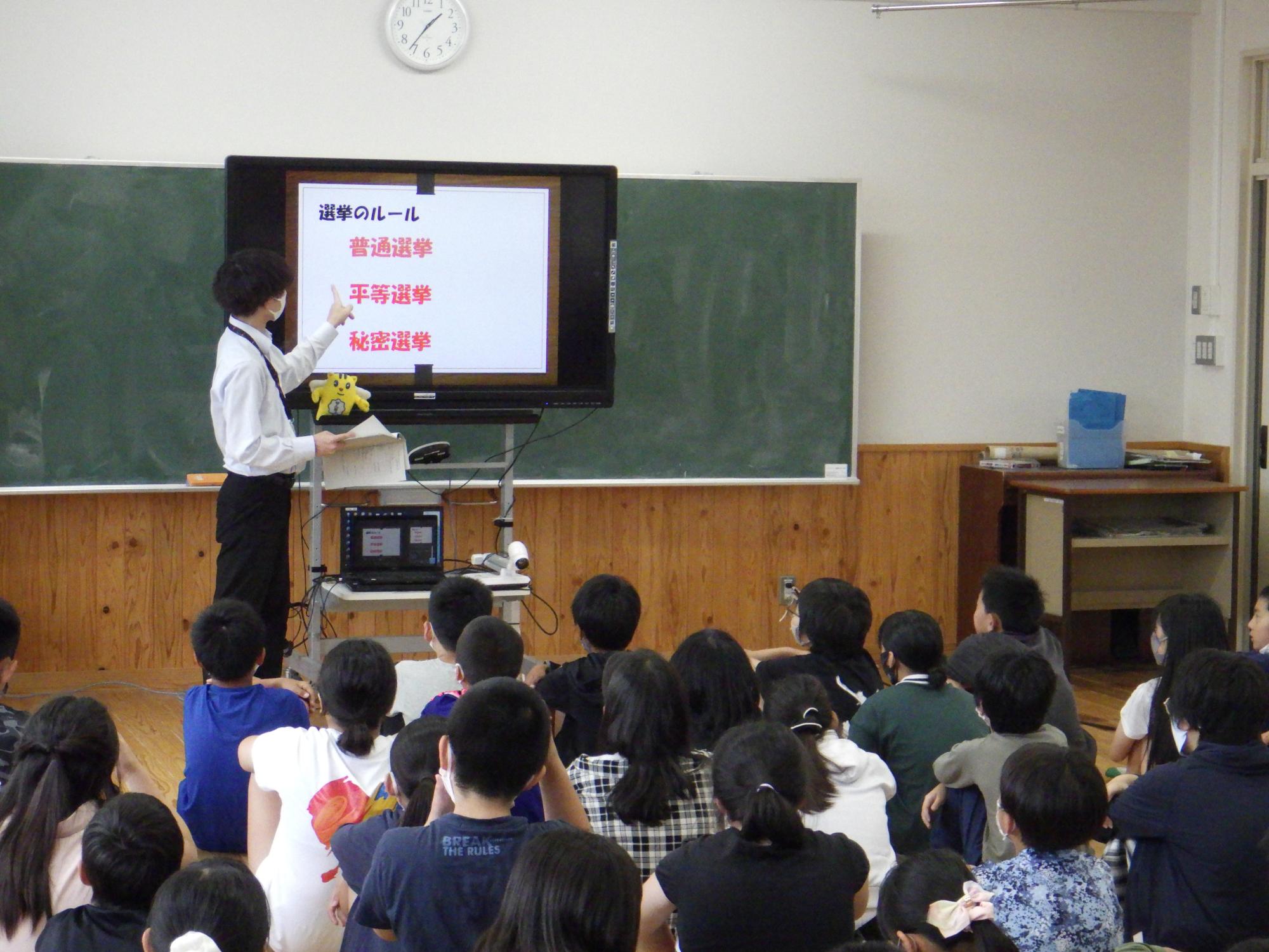 教室内で講師がモニターに映した「選挙のルール」という資料を指差しながら、普通選挙・平等選挙・秘密選挙について床に座った大勢の子供たちに説明している写真