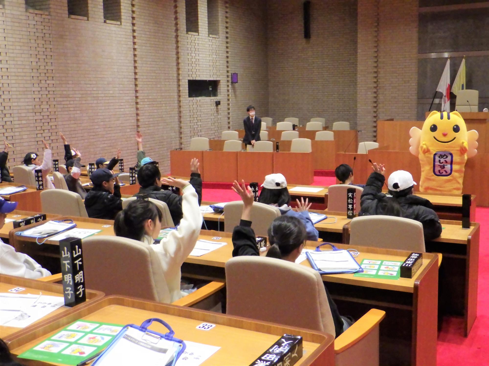 議場のような施設で、机の前に座った子供たちが一斉に挙手しており、前方にはスーツ姿の男性と黄色いキャラクターの着ぐるみ「めいすいくん」が立っている写真