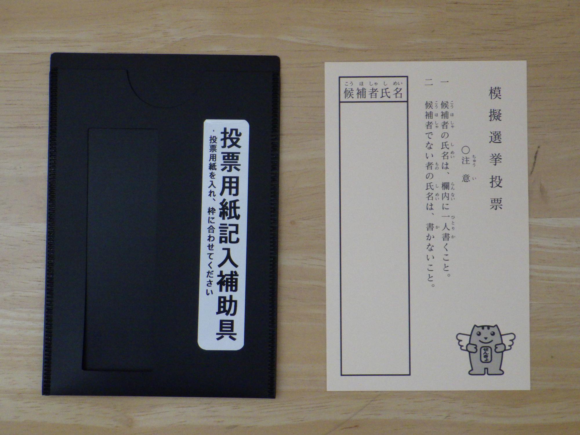 左側に「投票用紙記入補助具」と書かれた黒いファイル状の道具があり、右側に「模擬選挙投票」の用紙が並べて置かれている写真