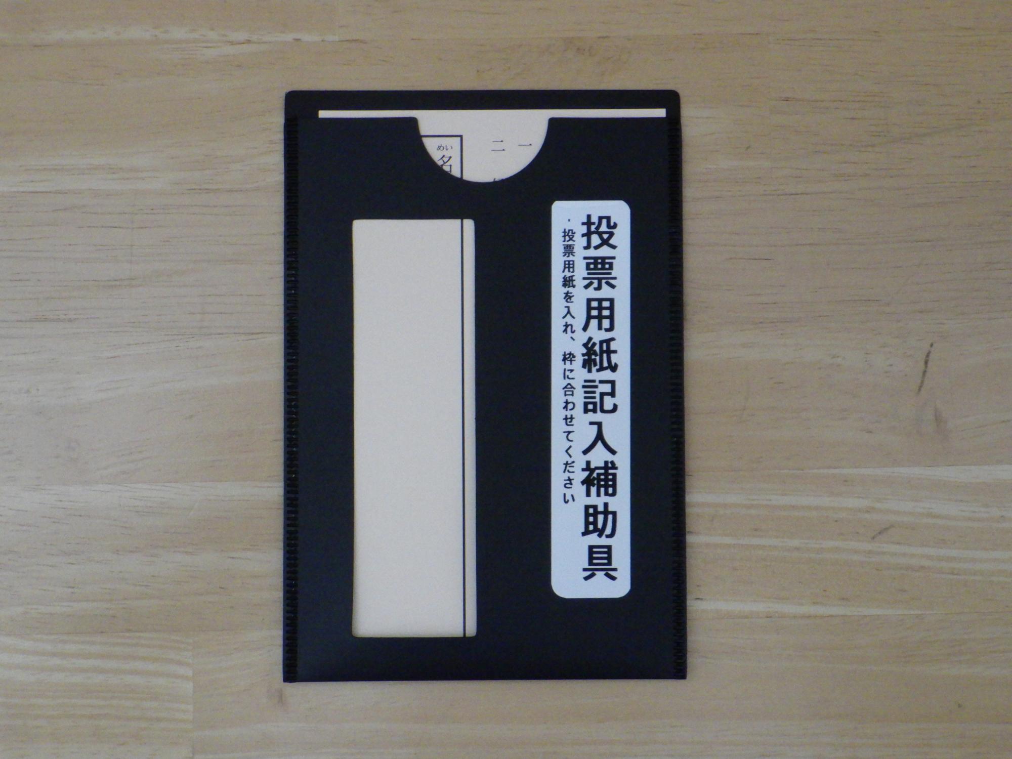 「投票用紙記入補助具」と記された黒いファイル状の道具が置かれ、その切り抜かれた長方形の窓から中に挿入された投票用紙の記入欄がちょうど見えるようになっている様子を俯瞰で撮影した写真