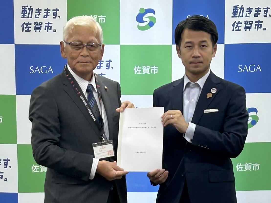 佐賀市のロゴと「動きます、佐賀市。」というキャッチフレーズが並ぶバックボードの前で、眼鏡をかけた大園会長と紺色のスーツを着た坂井市長が、一枚の協定書類を二人で持ちながら正面を見据えて並んでいる締結式での記念写真