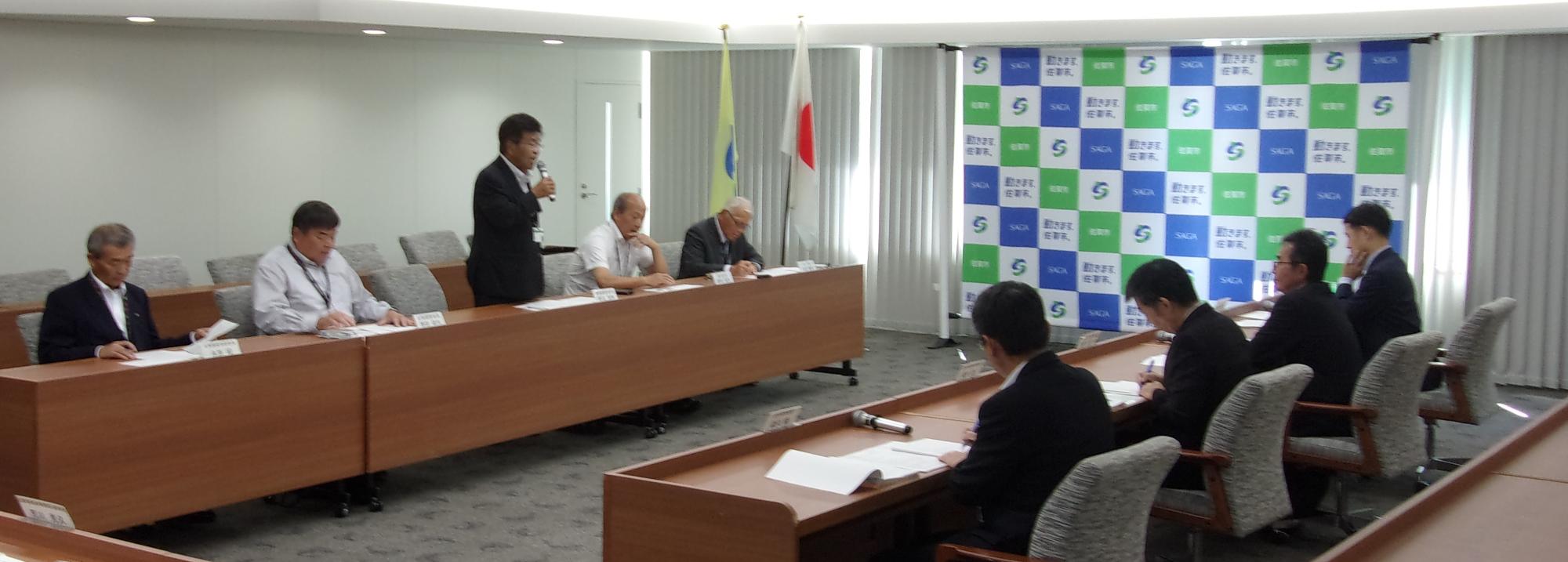 木目調の長い会議机が対面式に配置された室内で、数名の男性が席につき、マイクを持って発言する男性や手元の資料に目を落とす出席者たちの様子を捉えた、佐賀市の市章が描かれたバックボードが置かれた意見交換の様子の写真