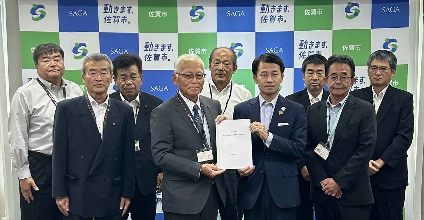 佐賀市のロゴと「動きます、佐賀市。」のキャッチフレーズが並ぶバックボードの前で、協定書を持つ中央の大園会長と坂井市長の周囲を、スーツやシャツ姿の関係者ら計9名が囲むようにして横一列に並んでいる締結式での集合写真