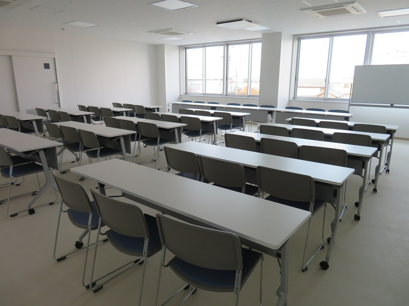 大きな窓際にホワイトボードが置かれ、整然と並んだ3人座りの白い長机とグレーの椅子がスクール形式で配置された産業振興会館 中会議室の写真