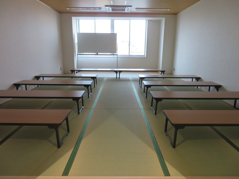 明るい光が差し込む大きな窓際にホワイトボードが置かれ、畳敷きの床に木目調の長机が左右対称に整然と並べられた産業振興会館 和室の写真