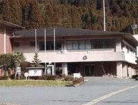 山林を背景に建てられたコンクリート造りの2階建ての建物で、正面には3本の掲揚ポールが立ち、入り口横の案内板や植栽が整えられた三瀬支所の外観写真