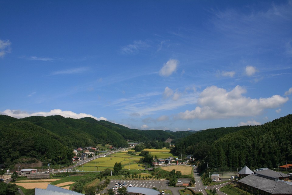 鮮やかな青空の下、豊かな緑の山々に囲まれた田園風景が広がり、手前には駐車場を備えた施設、中央には山あいを抜ける真っ直ぐな道路と点在する民家が見渡せる高台から写した写真