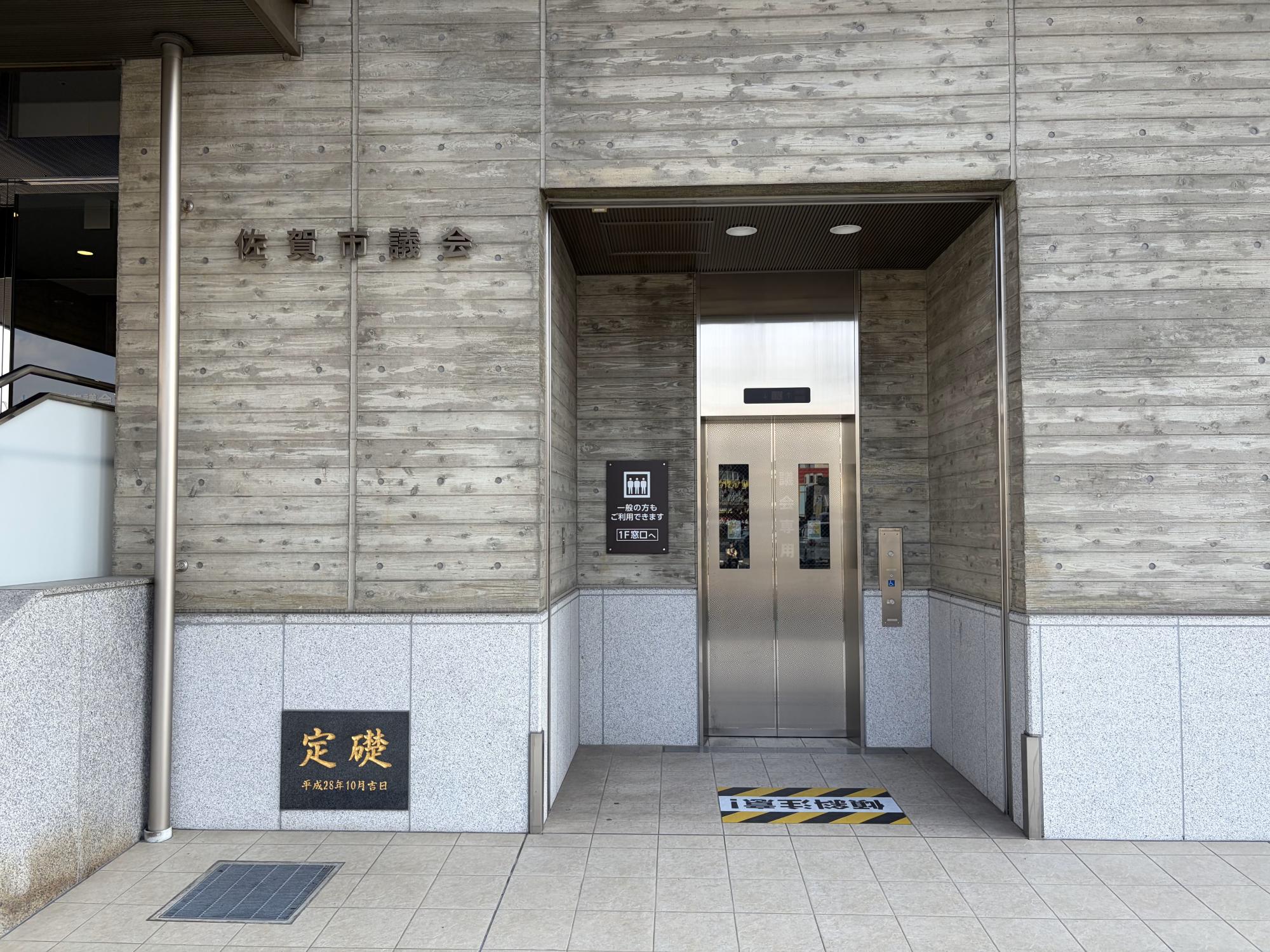 「佐賀市議会」の文字が壁面に掲げられた建物のエントランスで、木目調のコンクリート壁に埋め込まれたシルバーのエレベーターの写真