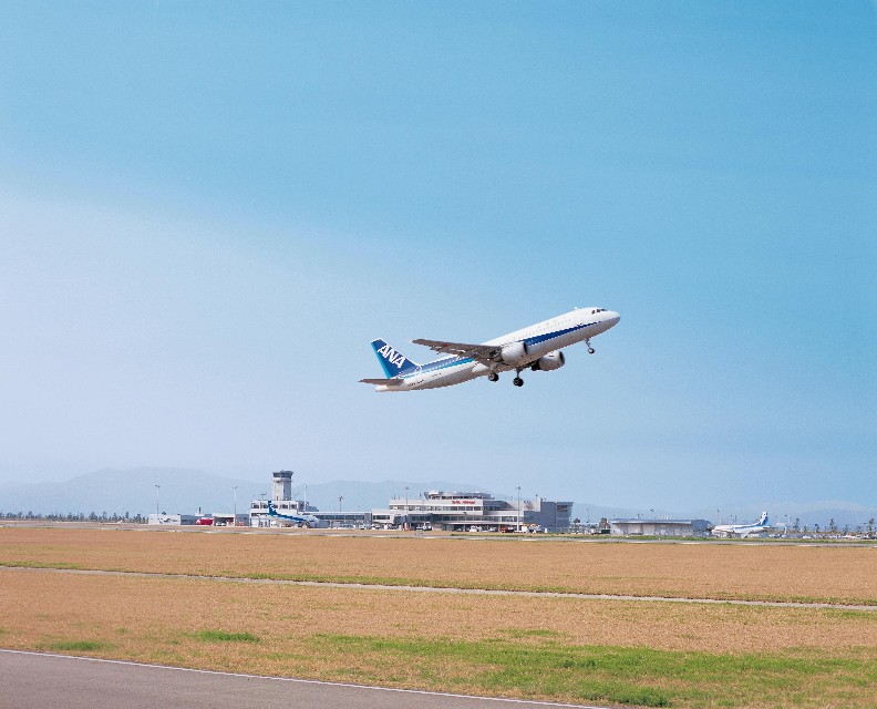 管制塔や空港施設が並ぶ広大な駐機場を背景に青空へと向かって、白い機体に青いライン入った旅客機が滑走路から力強く離陸する瞬間を写した佐賀空港の写真